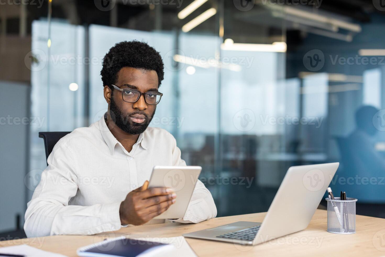 An African American man wearing glasses works at his office desk with a tablet and laptop. He is looking at the camera. photo