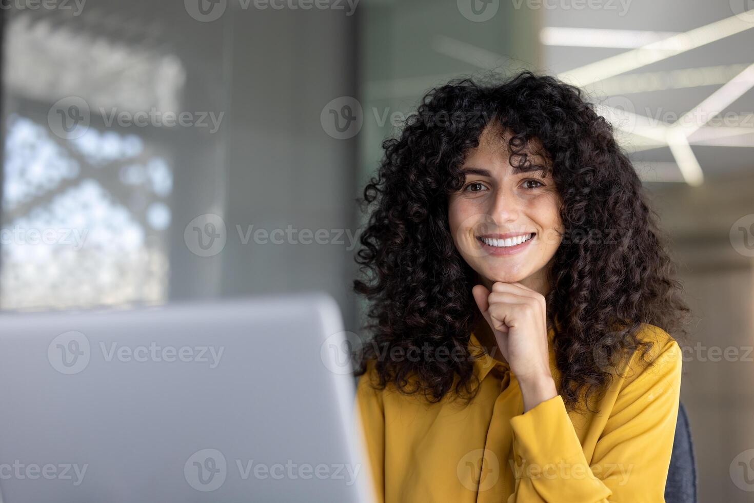 A smiling woman with curly hair rests her chin on her hand, working on a laptop in a bright, modern office setting photo