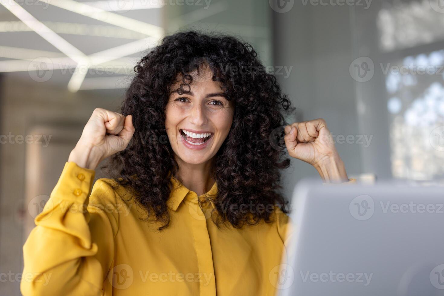 A joyful woman with curly hair celebrates success, raising her fists with an excited expression, in a bright setting. photo