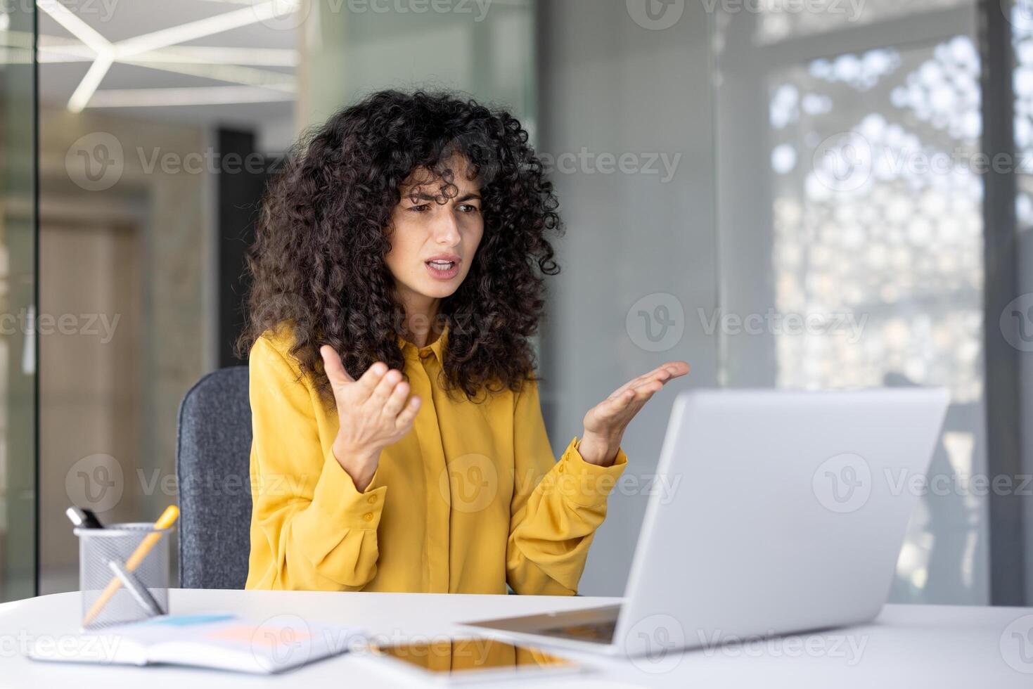 A woman with curly hair appears frustrated during a call, gesturing with her hands while seated in front of a laptop. photo