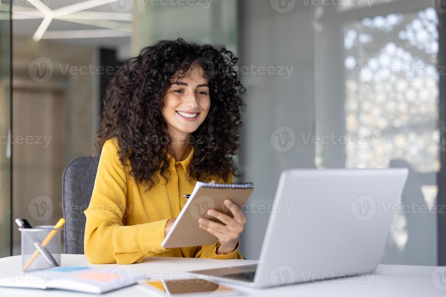 A smiling woman with curly hair takes notes while working on a laptop in a modern office setting. photo