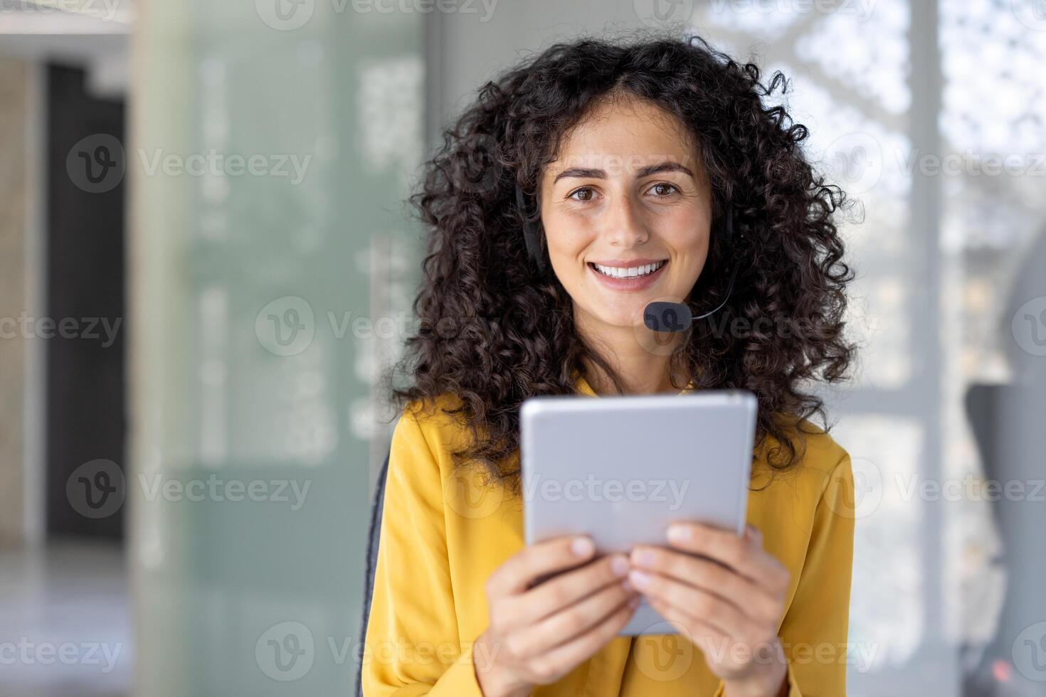 A smiling woman wearing a headset holds a tablet, appearing ready for customer service in a modern office setting. photo