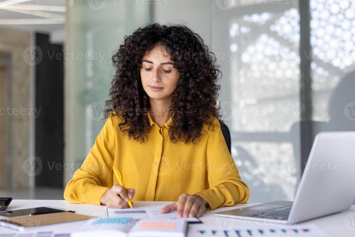 A businesswoman with curly hair is working at her desk, writing on a document with a pen in a modern office setting photo