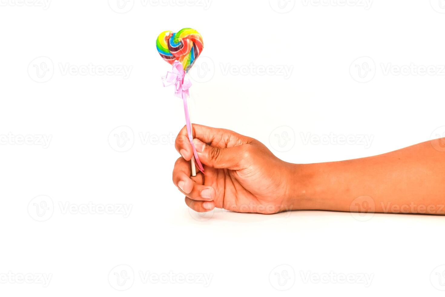 A child's hand holds a colorful heart-shaped lollipop against a white background, symbolizing sweetness and innocence photo