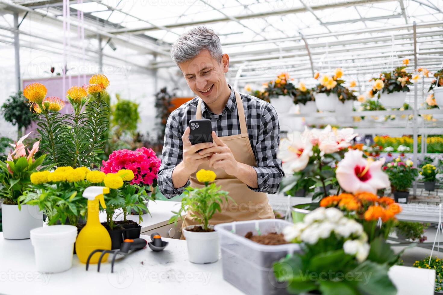 A smiling man in an apron uses his phone while standing in a vibrant greenhouse filled with a variety of colorful flowers and plants. photo
