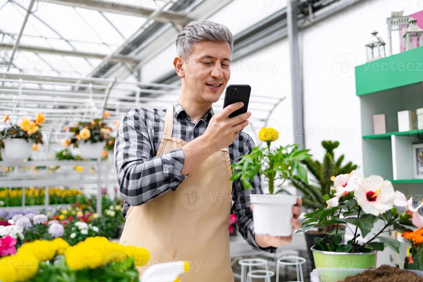 A man in a greenhouse takes a photo of a potted plant, surrounded by various flowers, using his phone.