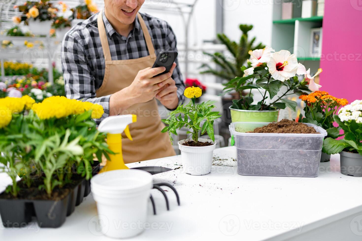 A florist taking a photo of a flower on a phone while surrounded by various plants and gardening tools.