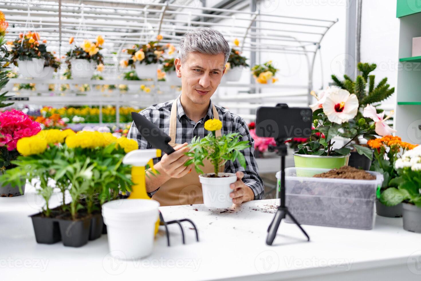 A man shows a plant while filming a in a greenhouse. Flowers and gardening tools are on a white table. photo