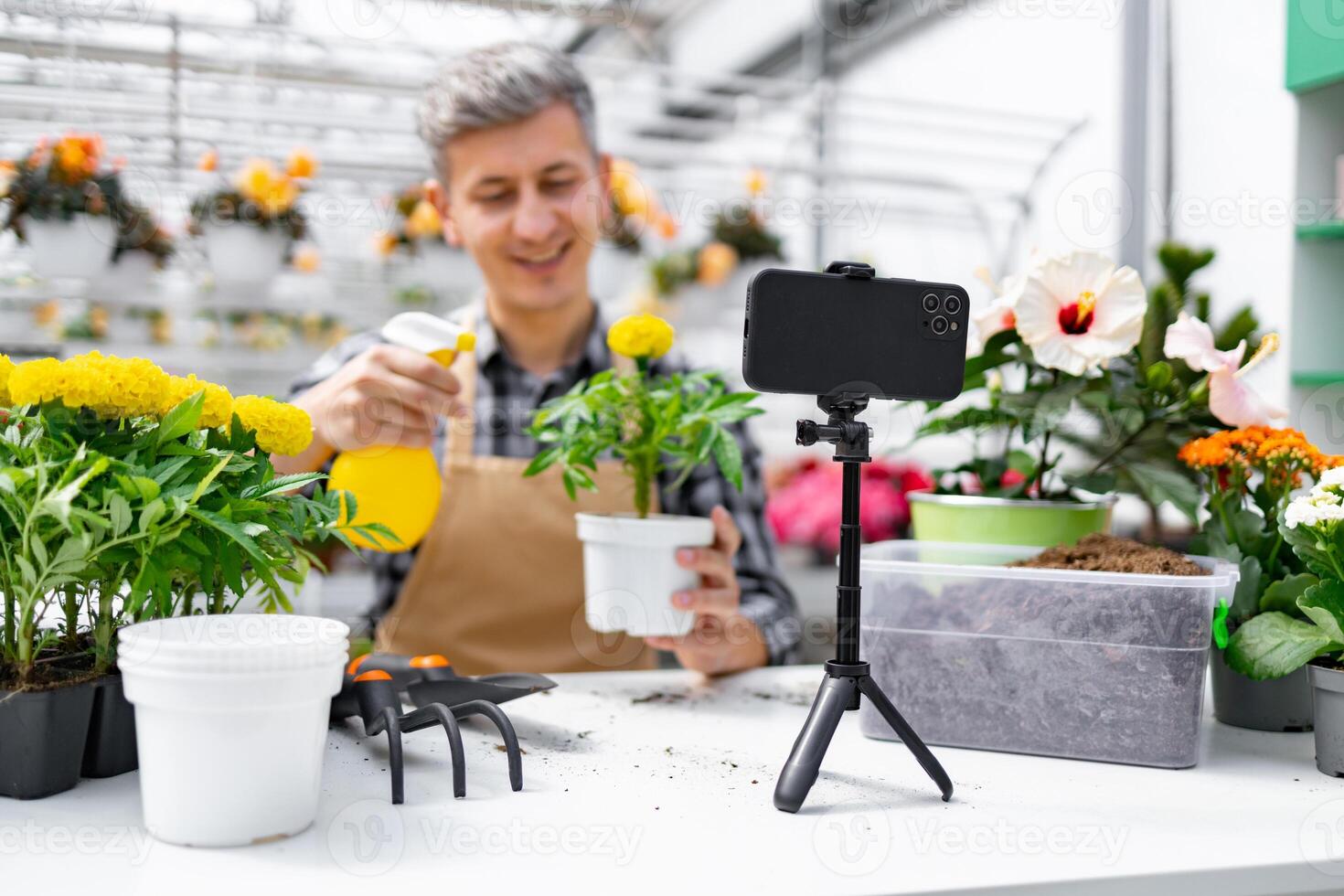 A man in a greenhouse records a gardening tutorial with his phone, surrounded by plants and flowers. photo