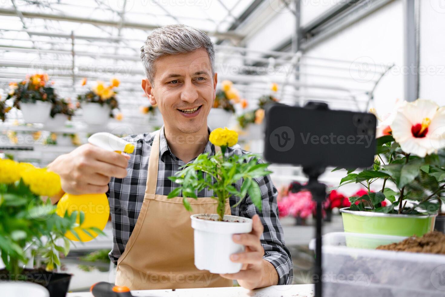 A cheerful man in a greenhouse demonstrates plant care, holding a sprayer and a potted marigold while recording a on his phone. photo