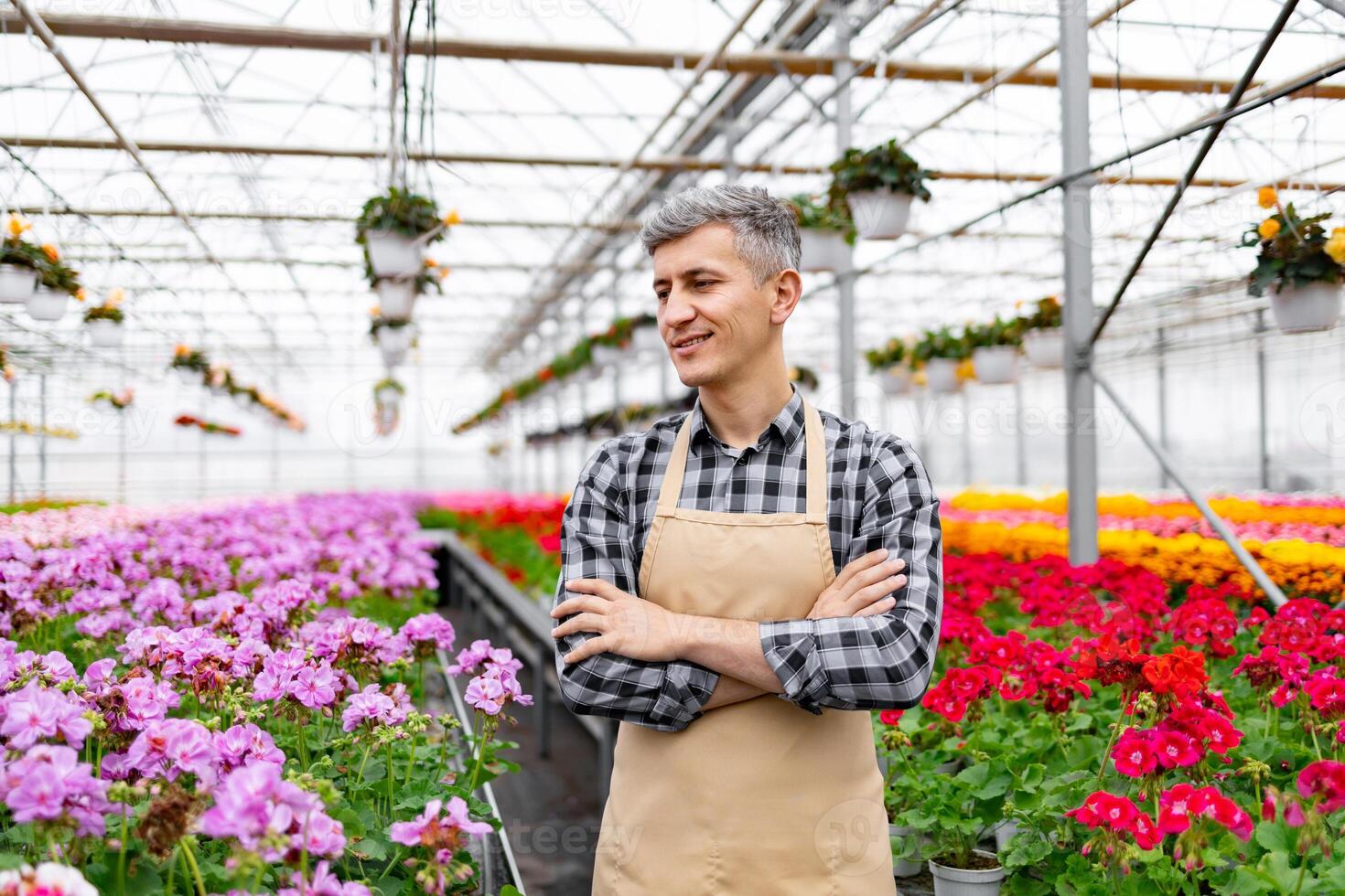 A smiling man with crossed arms stands in a sunlit greenhouse, surrounded by vibrant flowers and hanging plants. photo