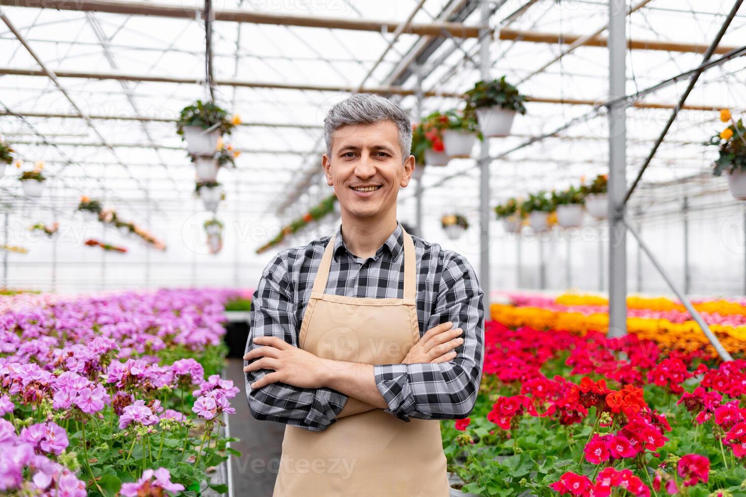 A smiling man in an apron stands in a greenhouse, surrounded by colorful flowers. photo