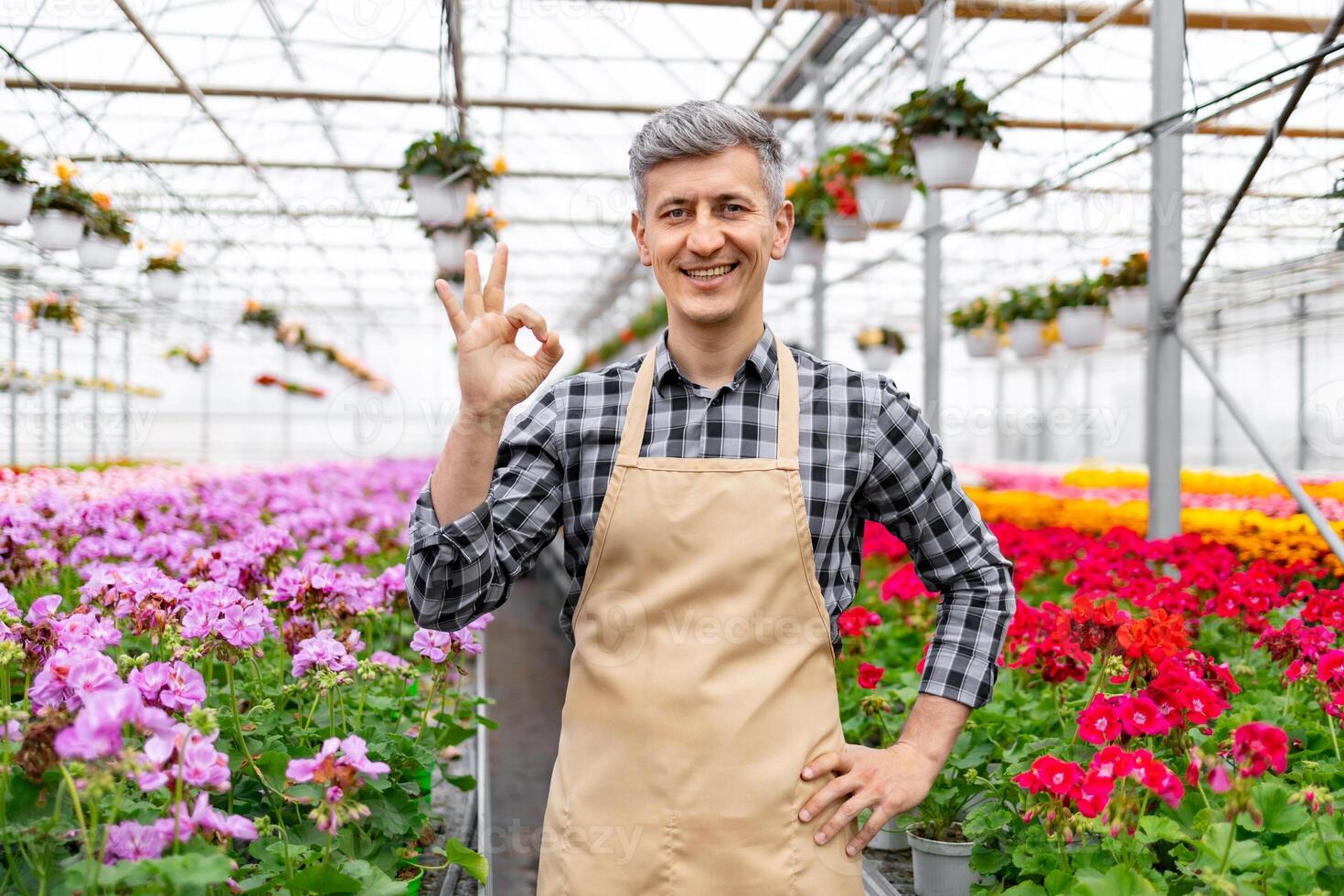 A friendly man in a greenhouse gives an 'okay' sign, surrounded by vibrant flowers, indicating everything is perfect. photo