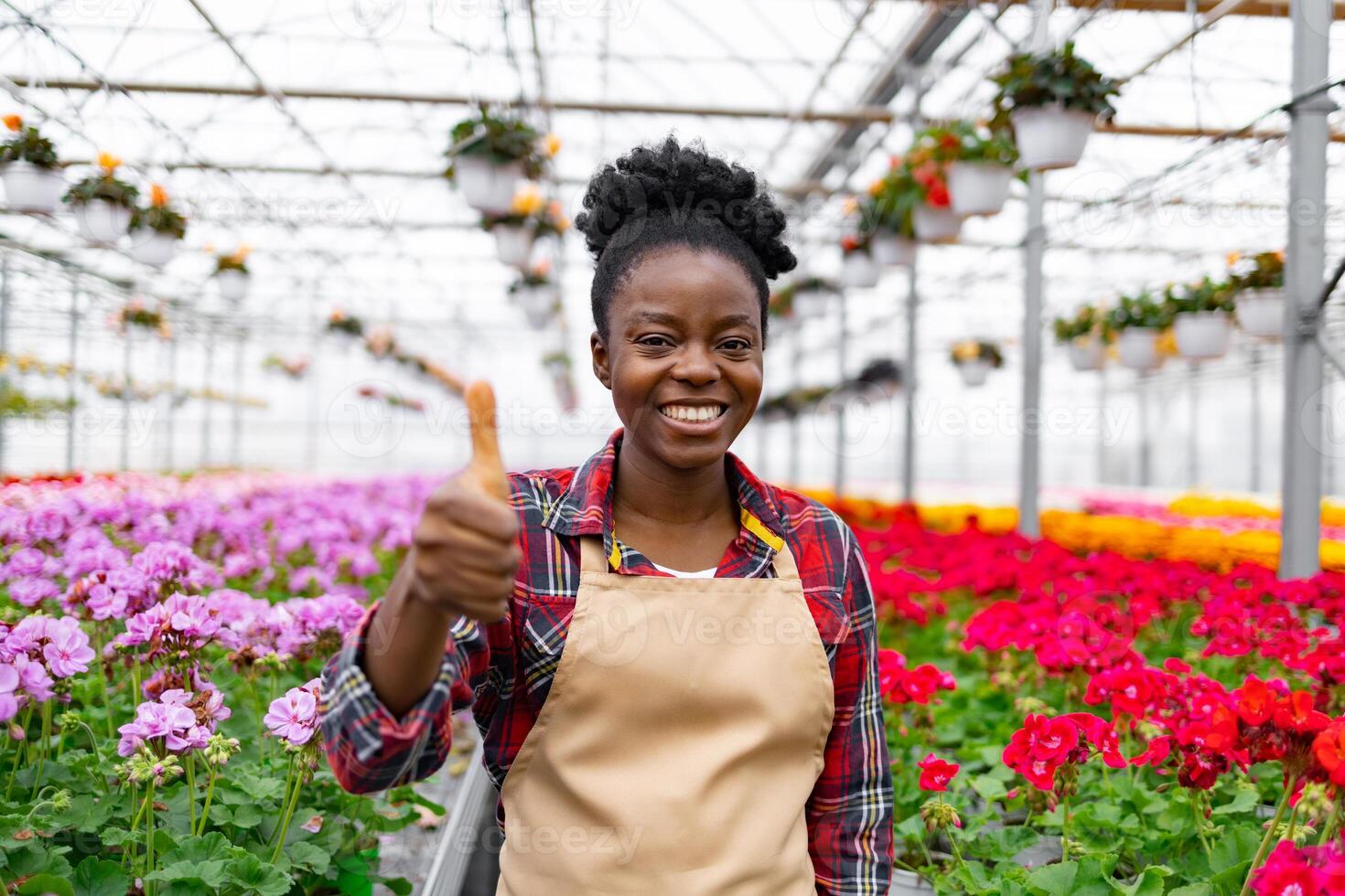 A smiling African woman with thumbs up in a greenhouse full of colorful flowers, showing approval of the thriving plants. photo