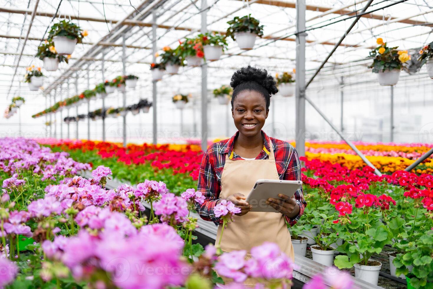 A smiling African woman in a greenhouse with a tablet, surrounded by vibrant flowers and plants, and other potted plants. photo