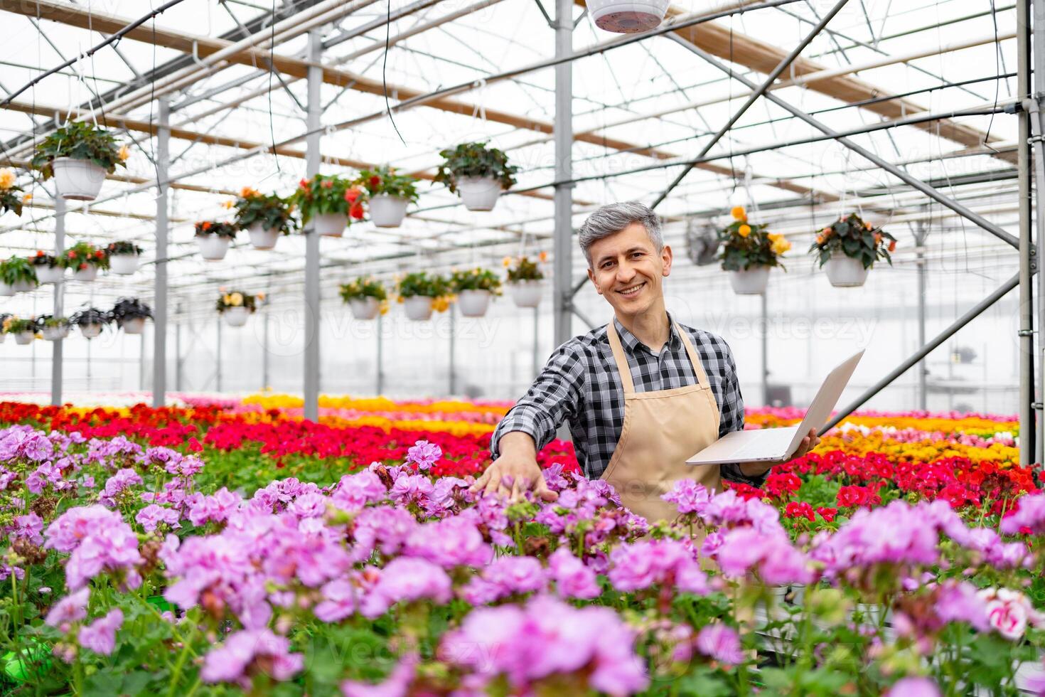 A smiling greenhouse worker examines flowers with a laptop, surrounded by vibrant blooms in a colorful floral arrangement. photo