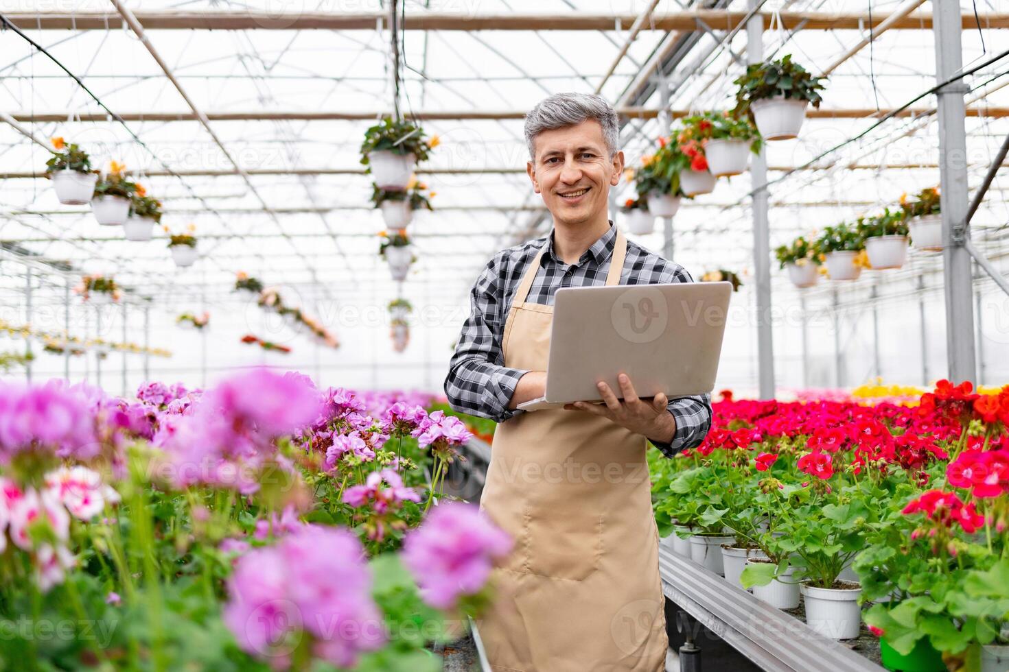 A smiling florist stands in a greenhouse, using a laptop to manage the plants. photo