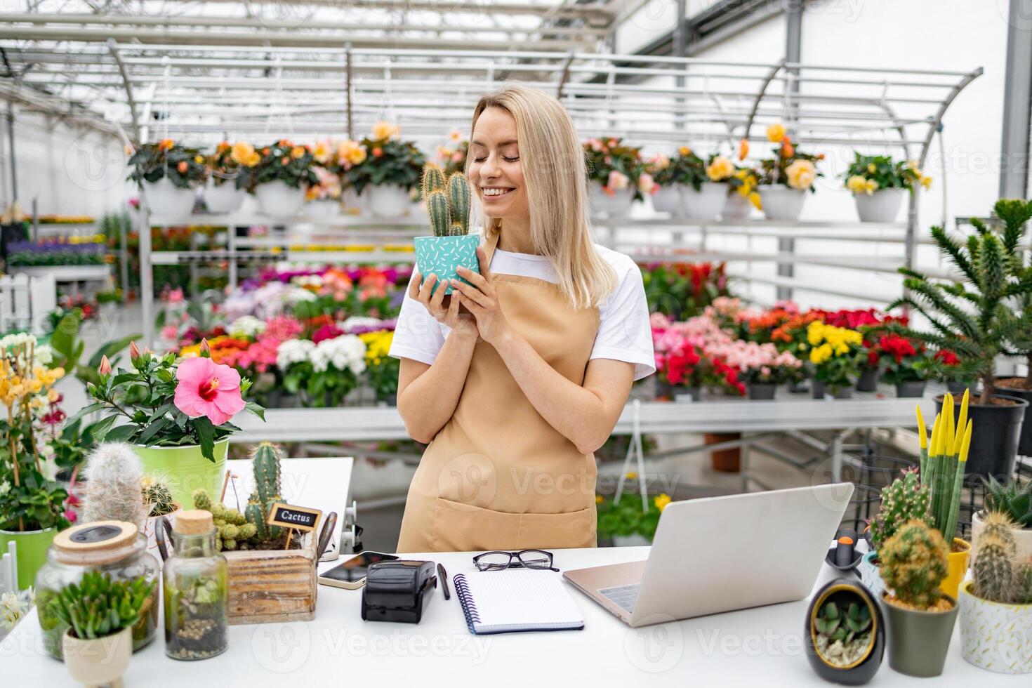 A smiling Caucasian woman, a florist, holding a cactus in her shop, next to a laptop and various plants for sale. photo