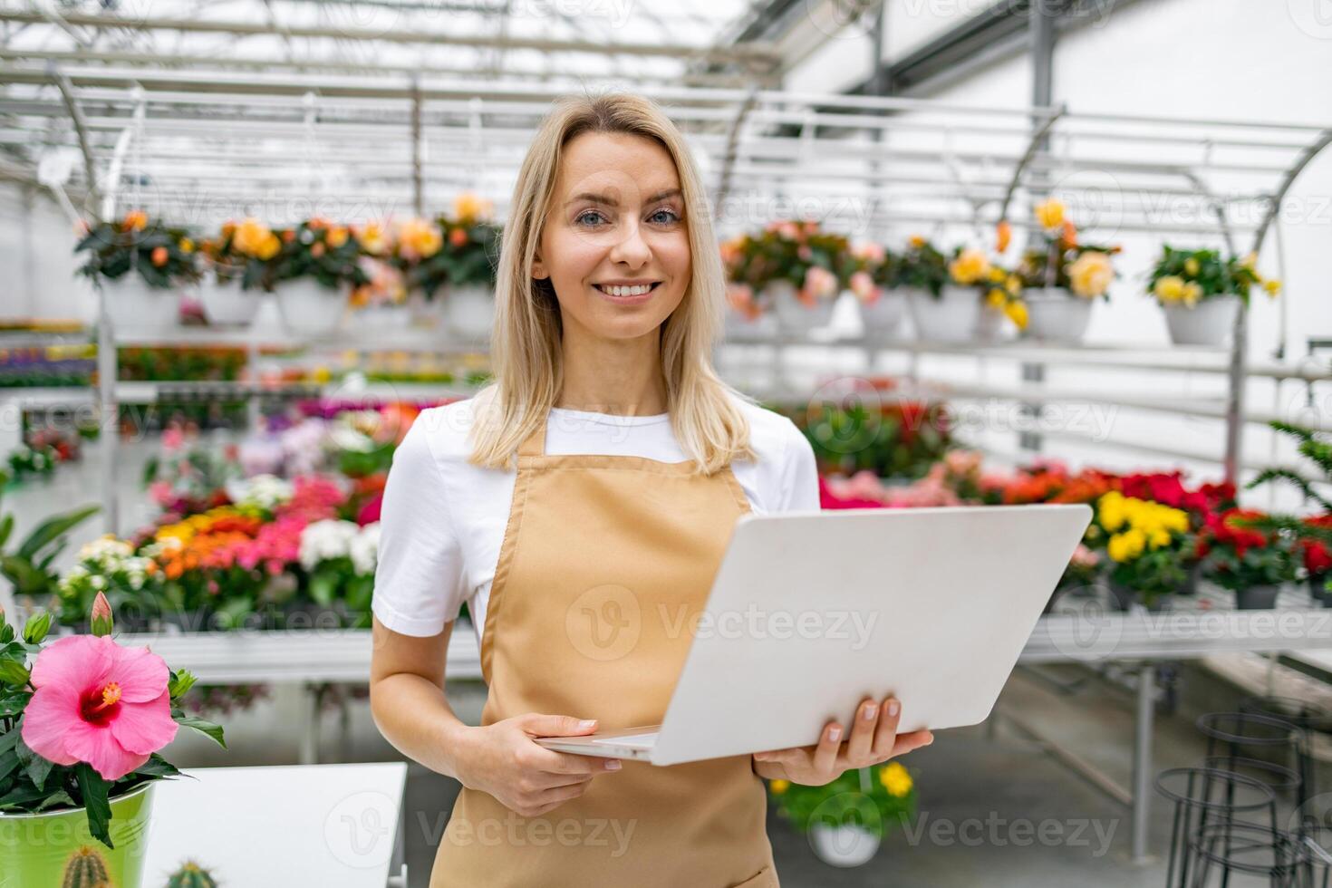 A smiling florist stands in her shop, holding a laptop, with an array of vibrant flowers in the background. photo