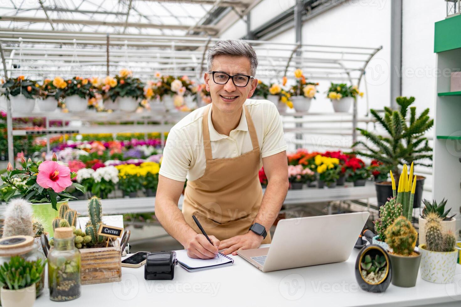 A Caucasian male florist writes in a notebook at a shop counter, surrounded by various plants and flowers. photo