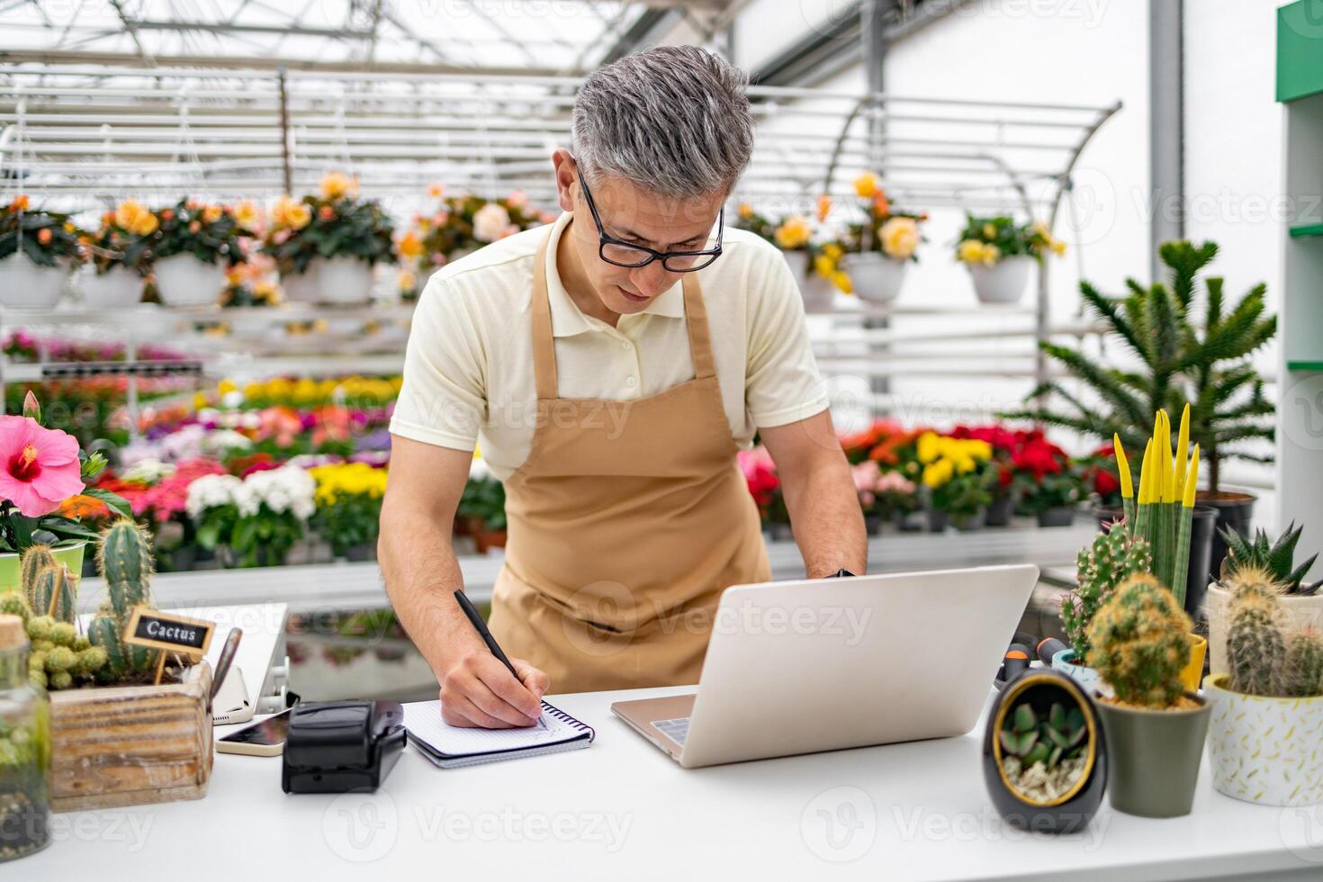 A Caucasian man, a florist, works on a laptop and takes notes at the shop's desk with various plants and flowers in the background. photo