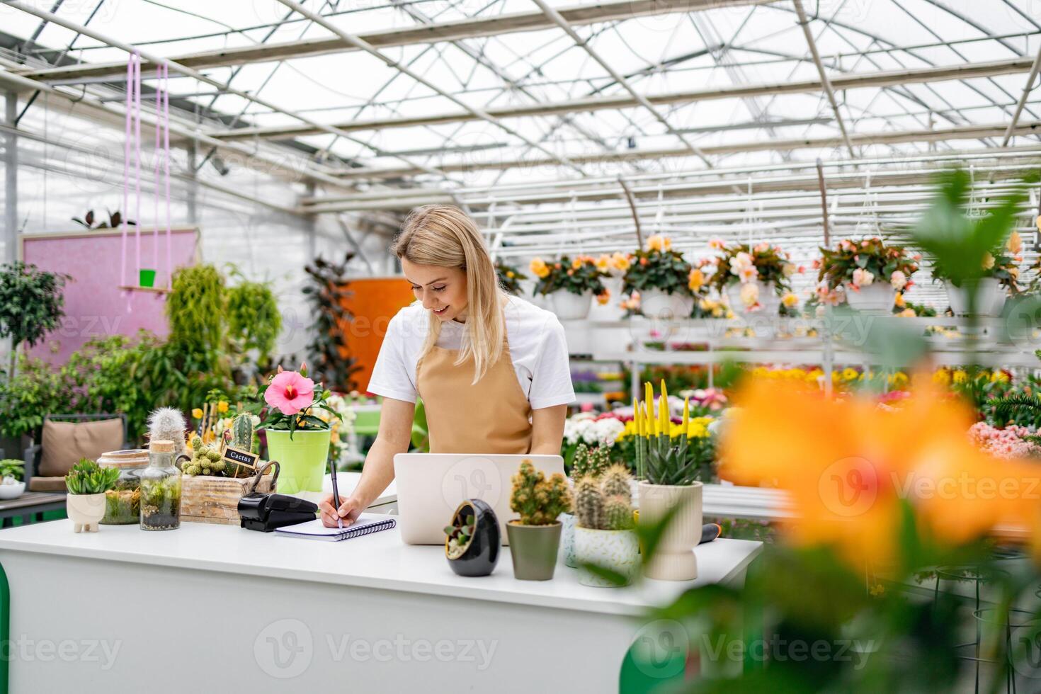 A Caucasian woman in a florist shop, taking notes at a counter, surrounded by various plants and flowers. She is smiling. photo