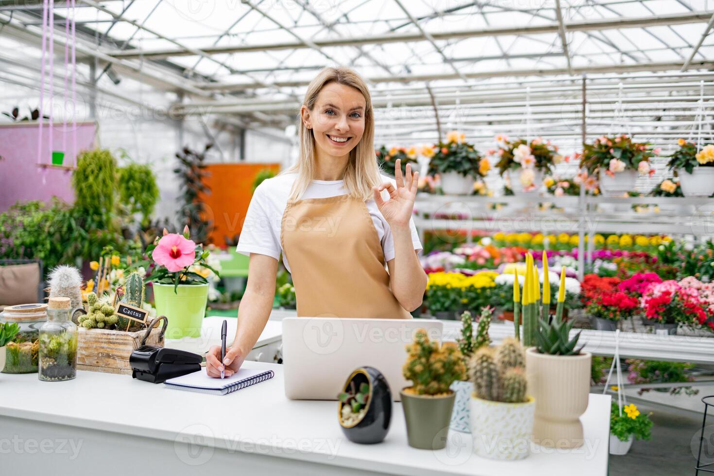 A smiling florist gestures with an 'OK' sign while standing at the sales counter, in her shop surrounded by plants and flowers. photo