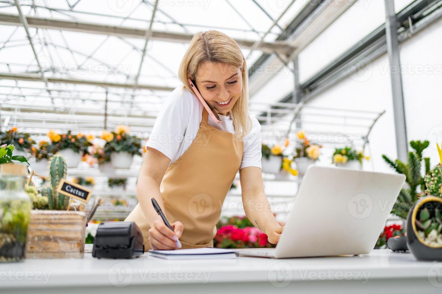 A Caucasian woman working in a florist shop, taking a call and using a laptop while writing. photo