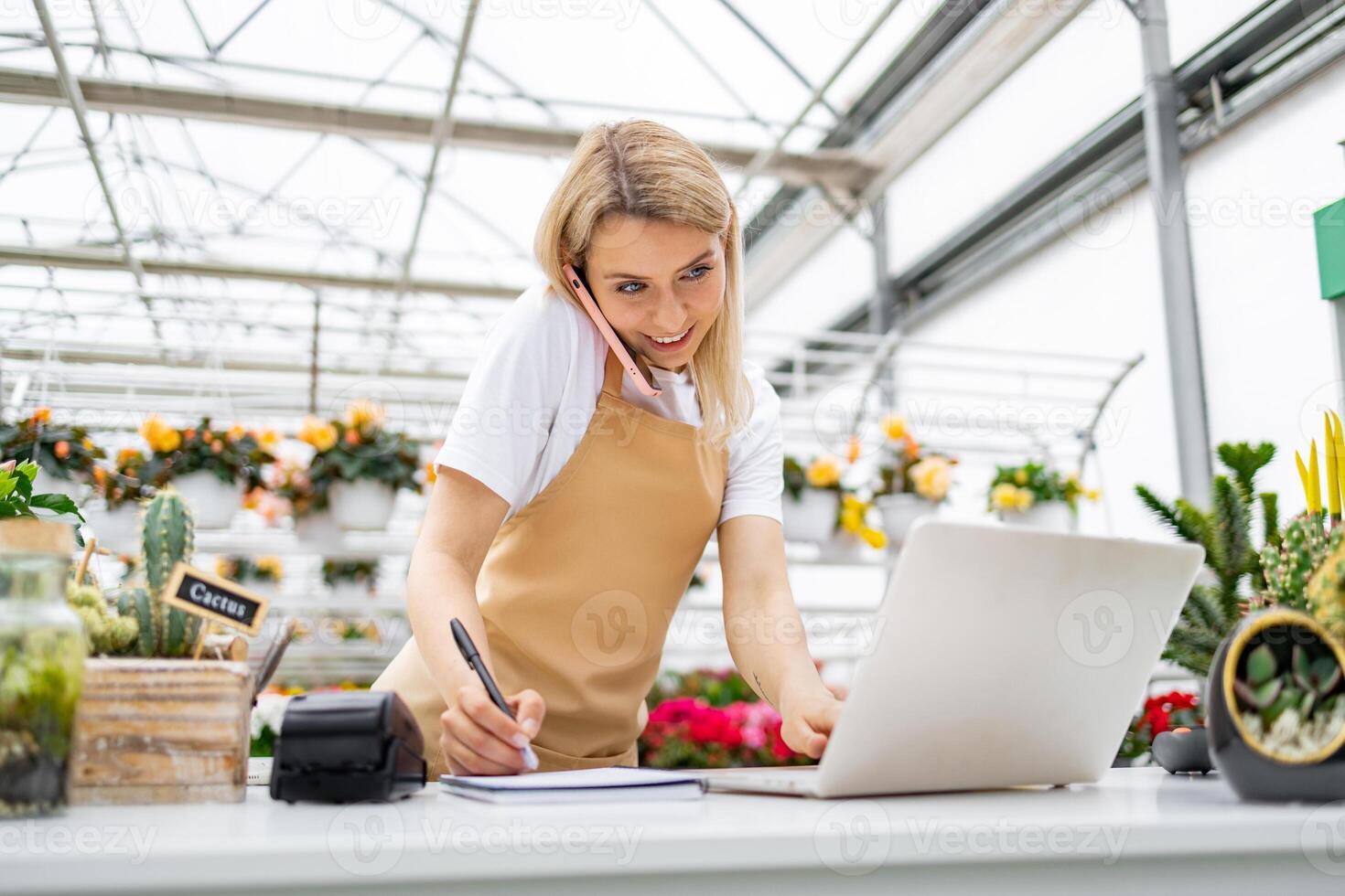 A Caucasian woman, working at a flower shop, is on the phone, taking notes, and using a laptop on a bright day. photo