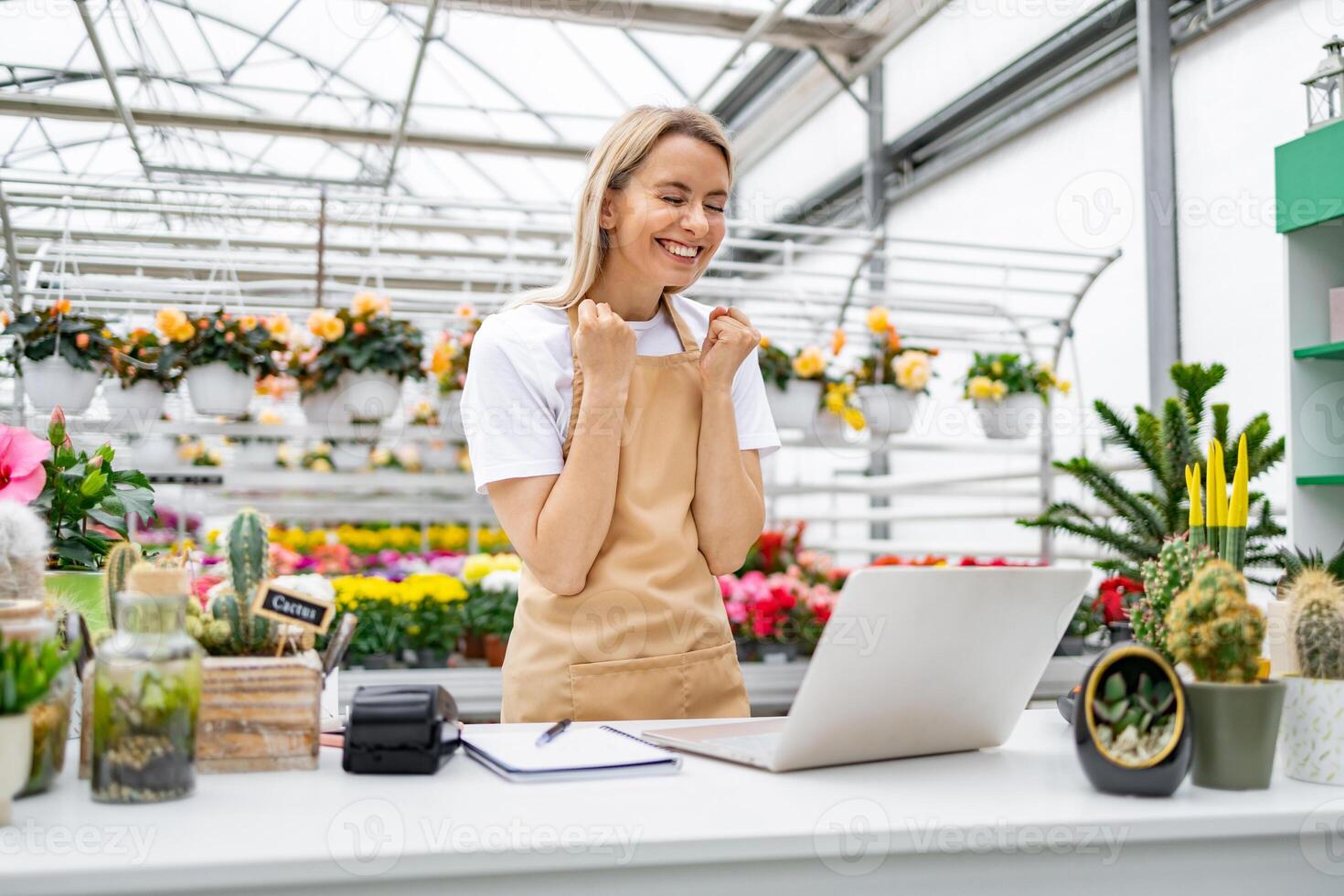 A joyful Caucasian woman in a florist shop celebrates a sale at the counter with a laptop, flowers, and plants around. photo