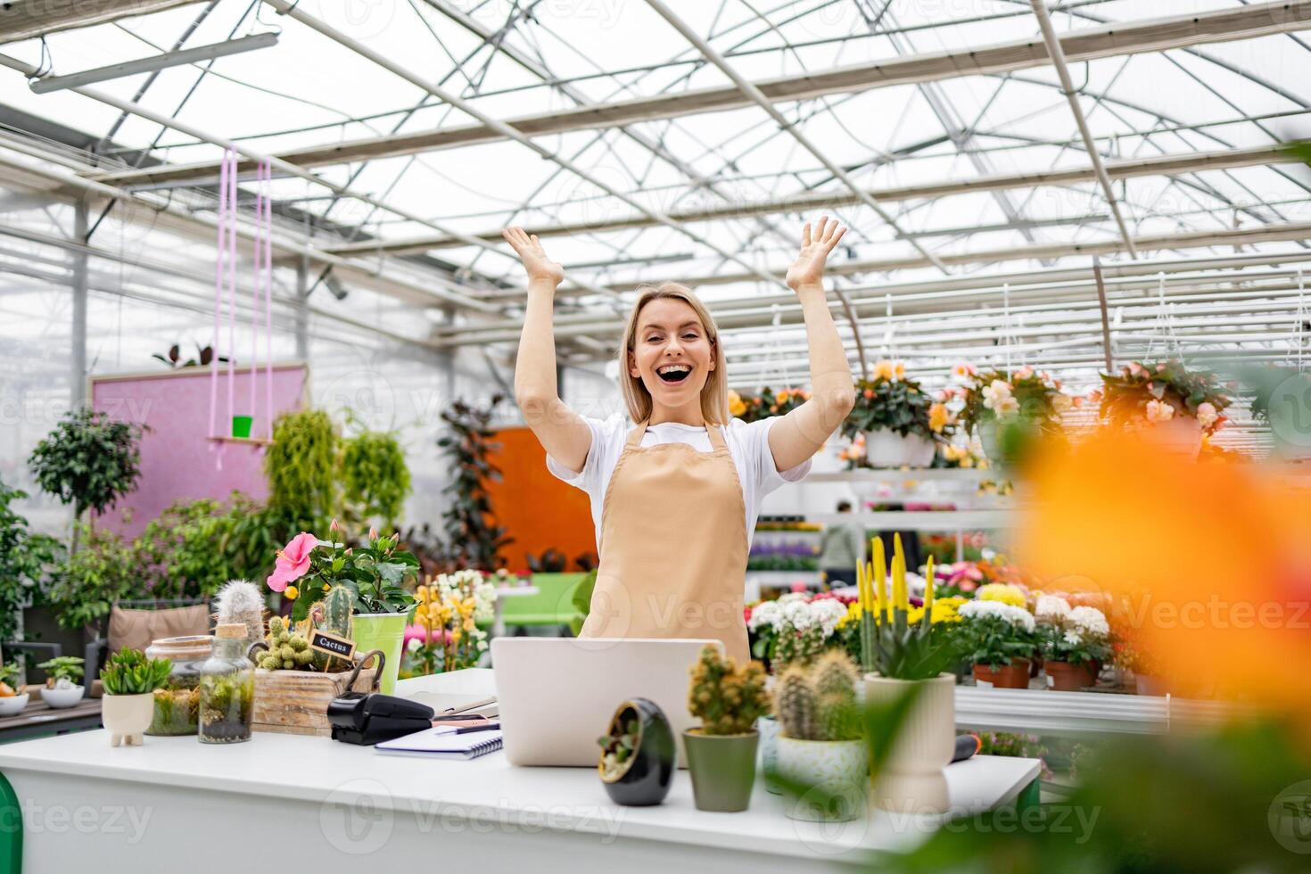 A Caucasian woman in a florist shop smiles with her arms raised as she works behind a counter filled with plants. photo