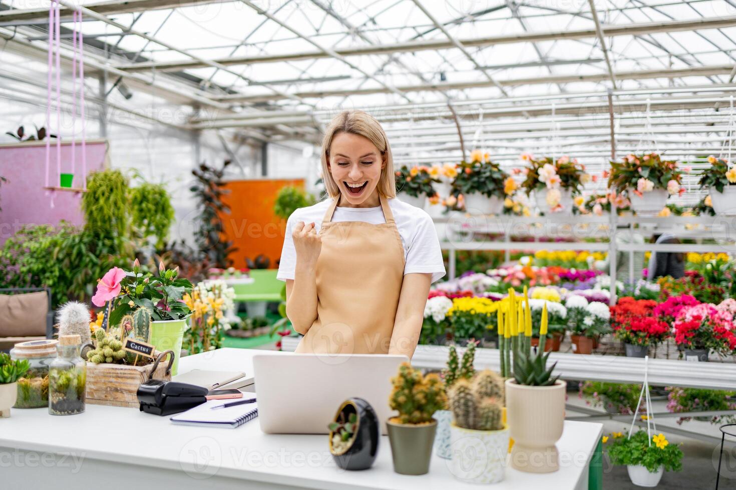 A happy Caucasian florist celebrates success at her shop using a laptop, surrounded by flowers and plants. photo