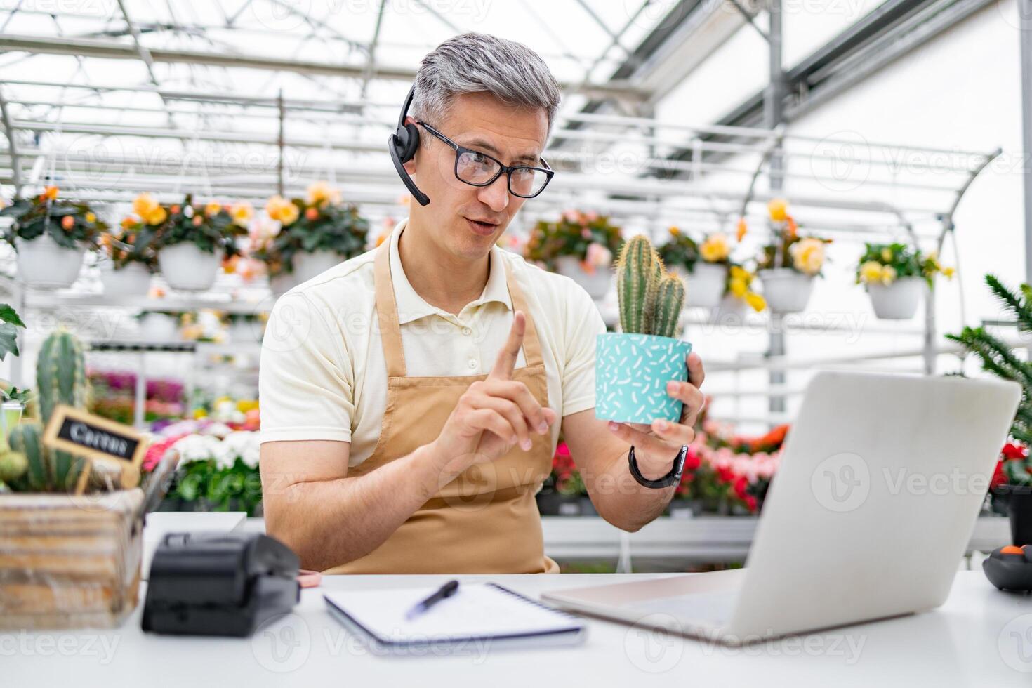 A Caucasian florist on a call presents a cactus while working from his flower shop, using a laptop to sell the plant. photo