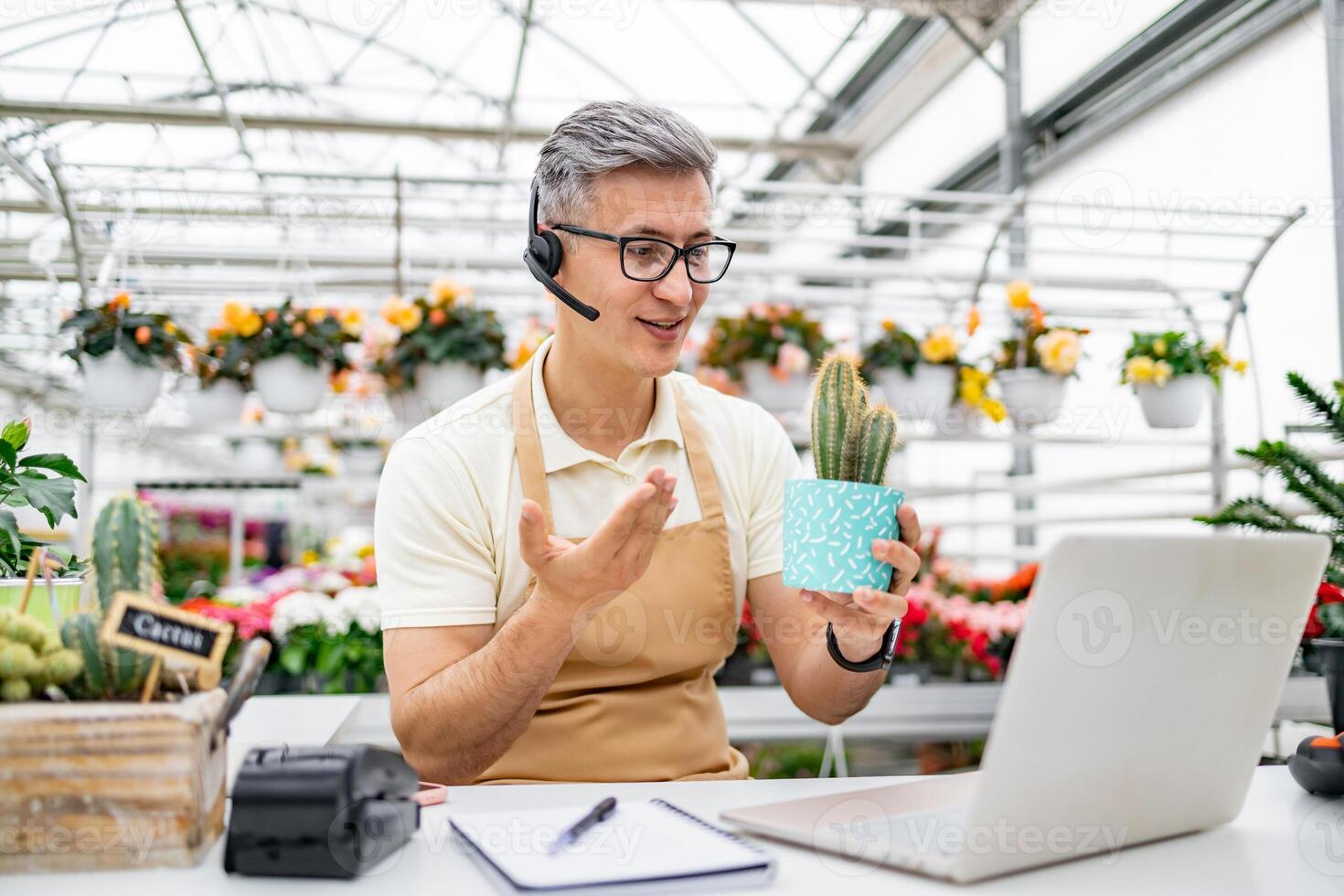 A Caucasian man in a flower shop uses a laptop while holding a potted cactus, presumably during a call for a sale. photo