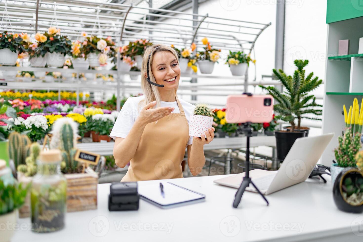 A Caucasian woman in a florist shop presents a cactus while live streaming for online sale from her table. photo