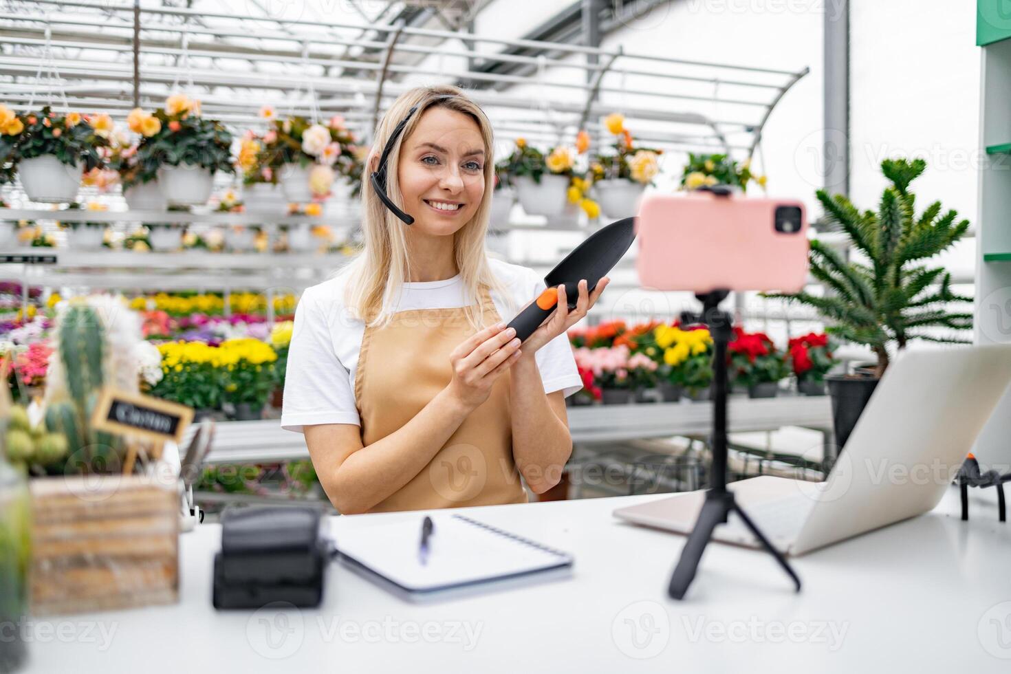 A Caucasian woman in a florist shop demonstrates a gardening tool, engaging in a live session with flowers in the background. photo