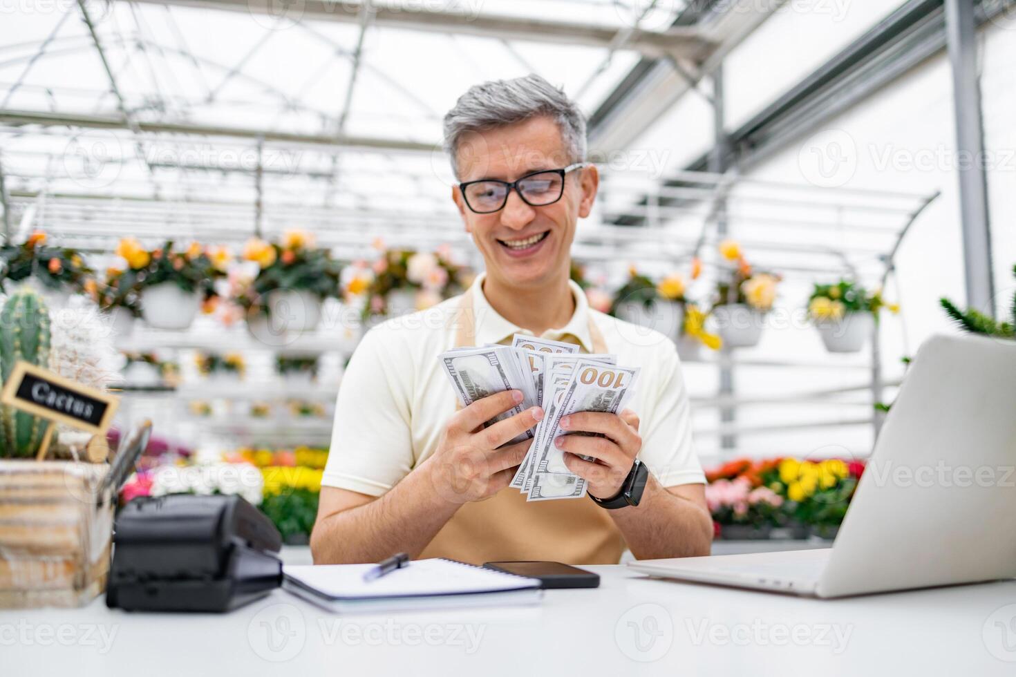 A Caucasian florist holds a stack of money at the shop's table, smiling with satisfaction at the sales. The shop has flowers. photo