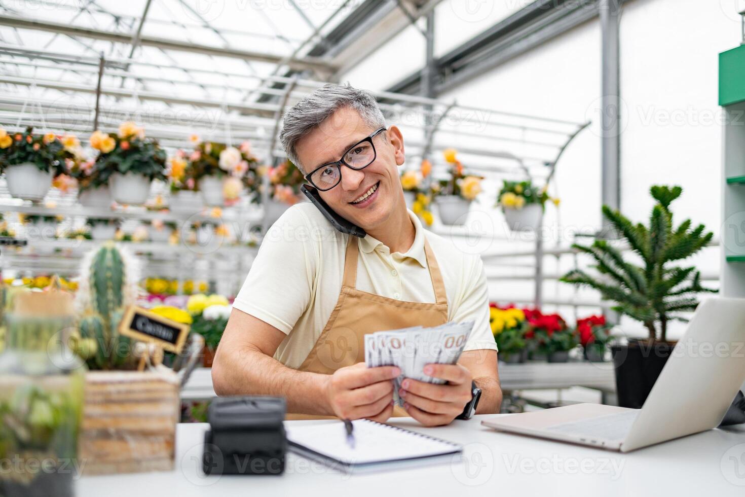 A Caucasian florist, happy with money in hand, smiles while on a phone call in his shop, surrounded by plants and flowers. photo