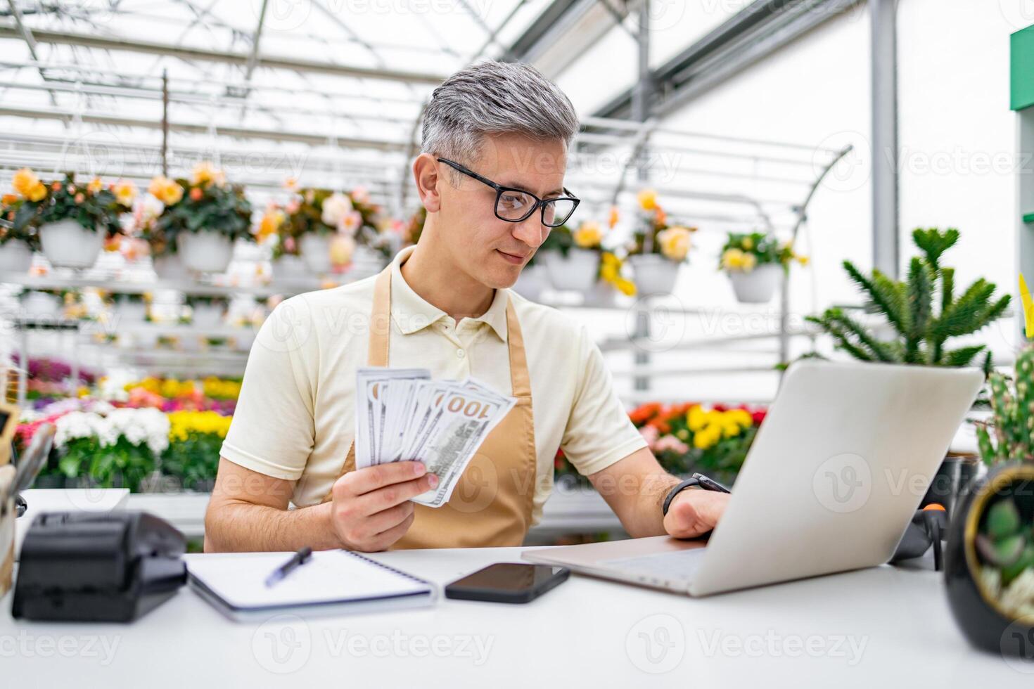 A Caucasian florist, holding cash, uses a laptop at a table in a flower shop, managing sales and finances. photo