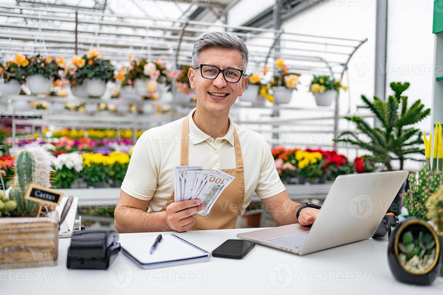 A smiling Caucasian man holds money, works on a laptop at a flower shop, surrounded by beautiful plants. photo