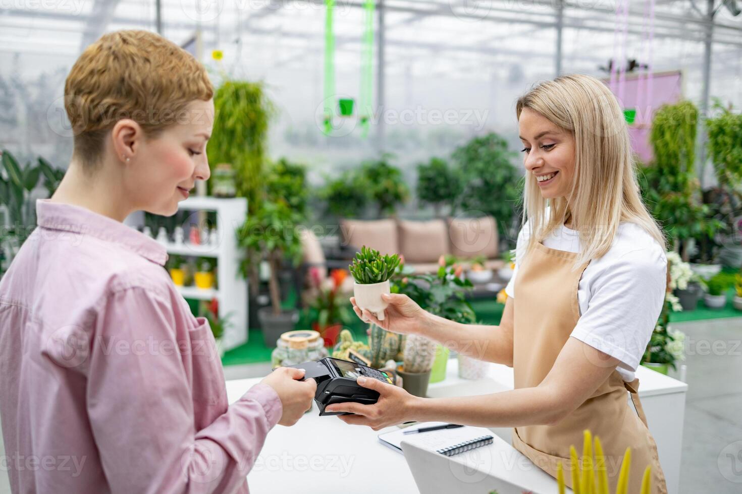 A smiling florist hands a small potted plant to a customer who is paying with a credit card. The sale is taking place in a plant shop. photo