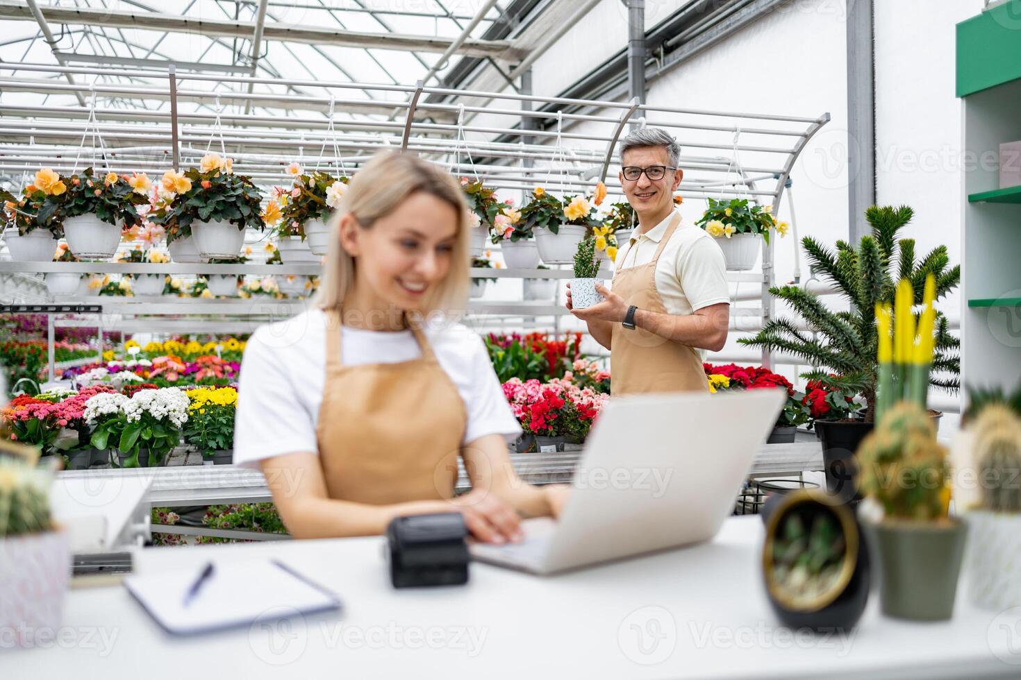 A Caucasian florist and seller happily working at their shop's checkout. Plants fill the background, and a man poses with a potted plant. photo