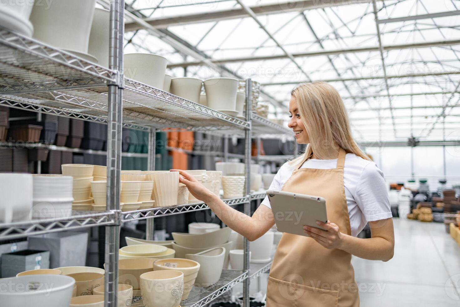 A woman in an apron looks at flowerpots in a shop. She is holding a tablet. photo