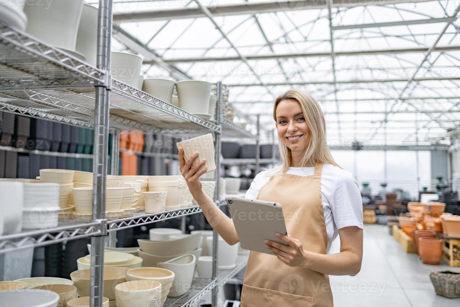 A smiling Caucasian woman holding a flowerpot and tablet inside a plant shop. Shelves are filled with various plant pots. photo
