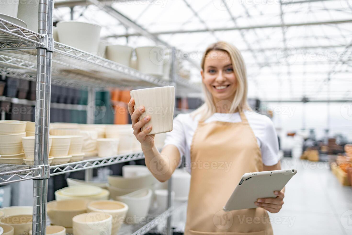 A cheerful woman in an apron holds up a flowerpot in a plant shop, with shelves of pots in the background. photo
