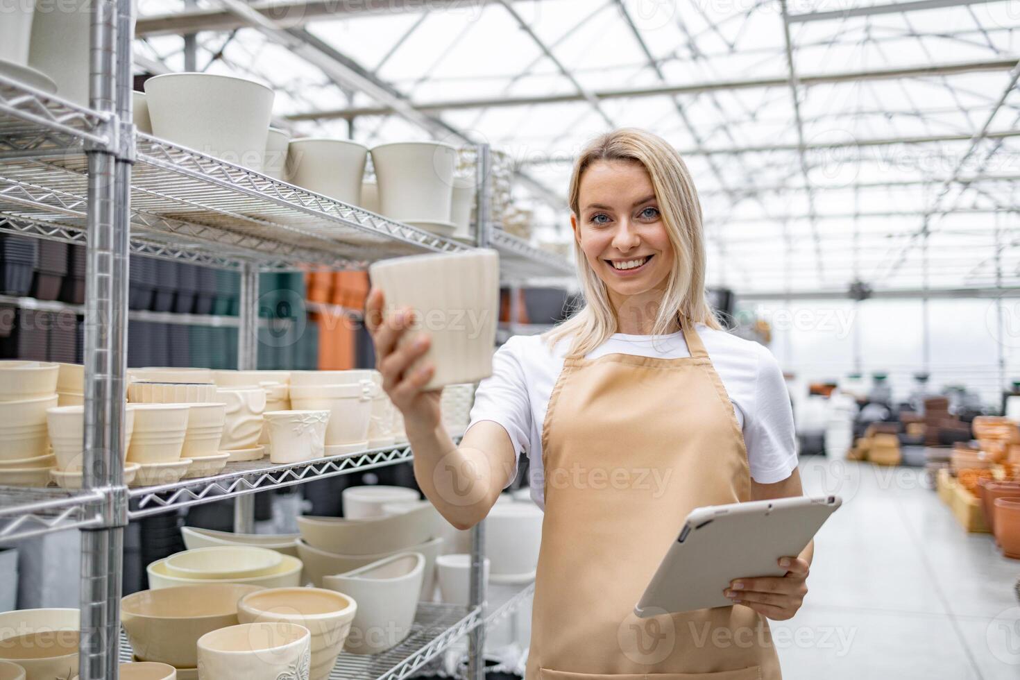 A blonde Caucasian woman in an apron selects a flowerpot in a shop. She is holding a tablet, smiling at the viewer, showcasing various plant containers. photo