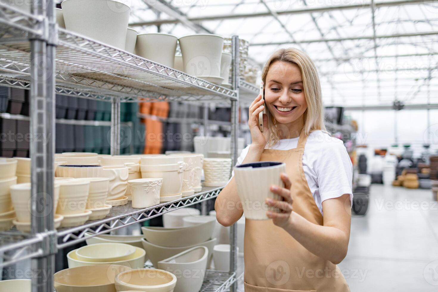 A Caucasian woman examines a flowerpot while talking on her phone inside a plant shop. Various other flowerpots are on shelves in the background. photo