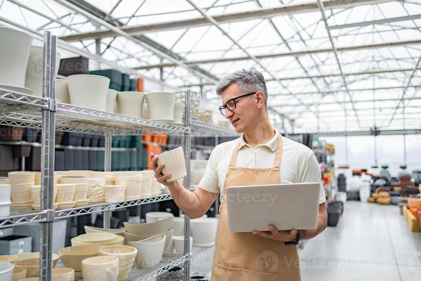 A Caucasian man inspects a flowerpot in a plant shop, holding a laptop for inventory. The shop is organized with shelves full of various pots. photo