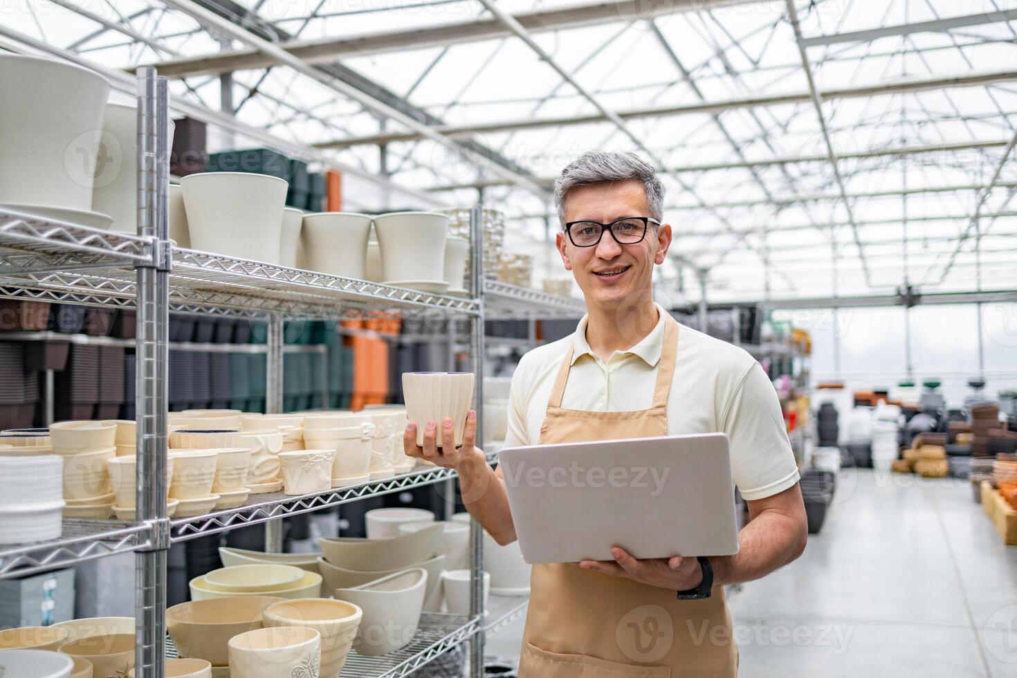 A caucasian man in glasses holds a flowerpot and a laptop in a shop full of flowerpots. photo