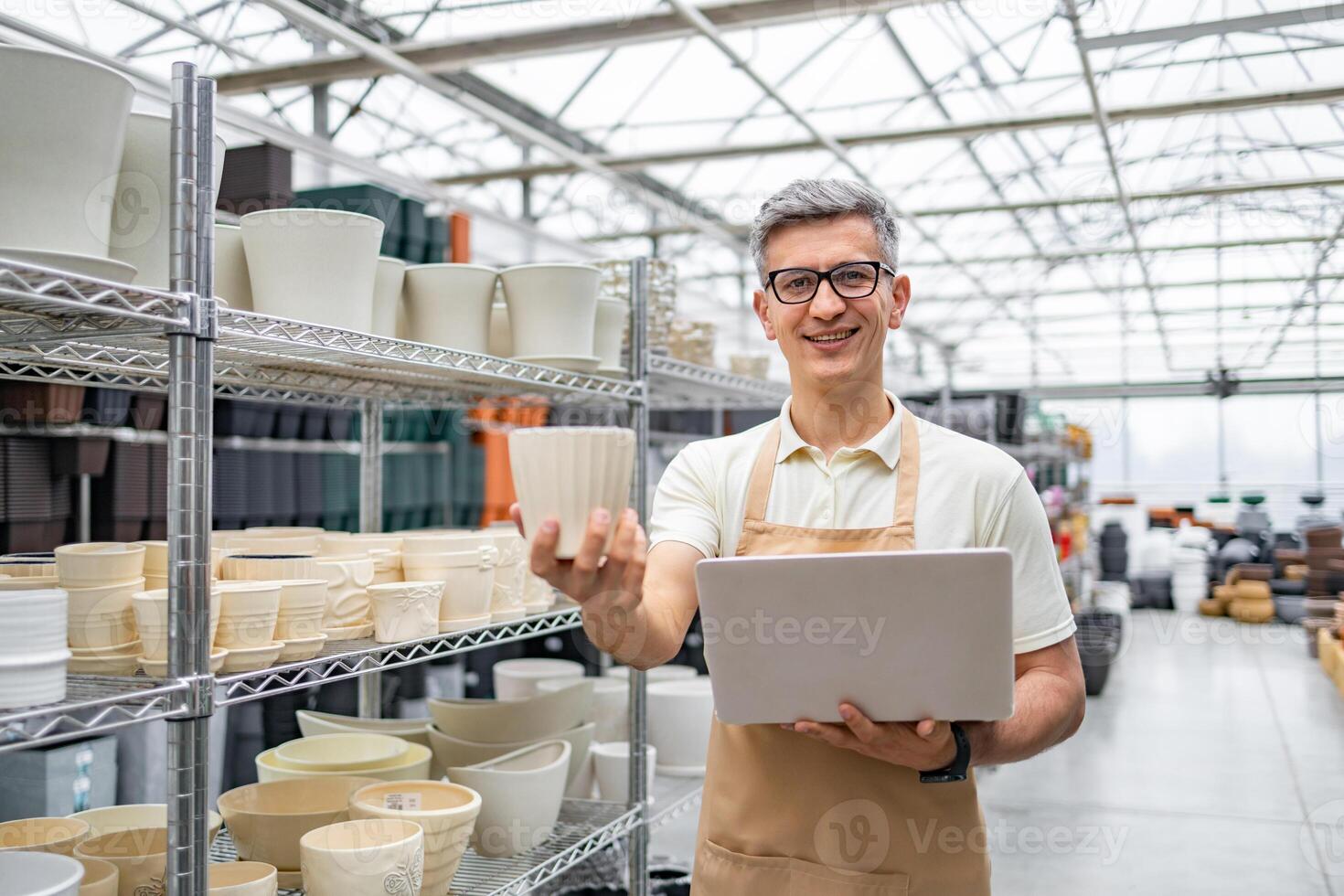 A smiling man in a plant shop holds a flowerpot and a laptop, with shelves of other pots behind him. photo